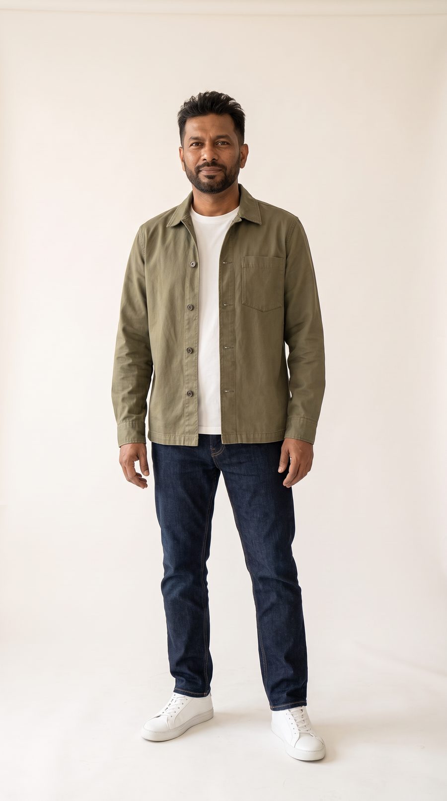 The same man: textured cut, short boxed beard, olive overshirt open over a white tee, dark jeans, white sneakers